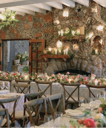 A long table dining table with floral arrangements next to a stone fireplace